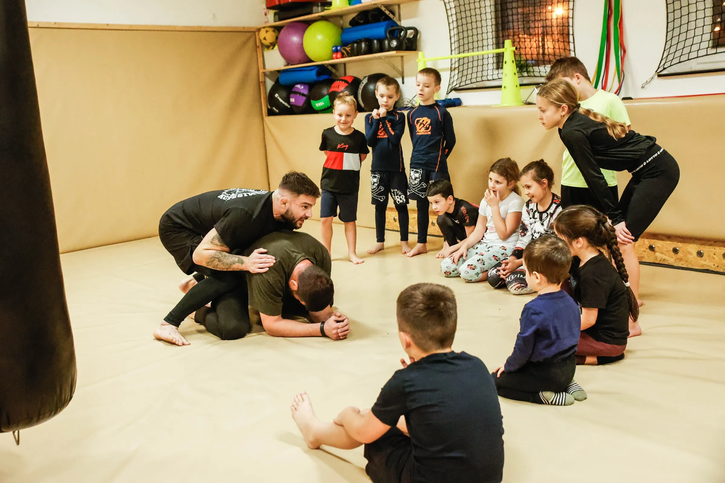 Trenér ukazuje techniku na zemi skupině dětí, které sedí kolem něj na tatami a sledují ho.