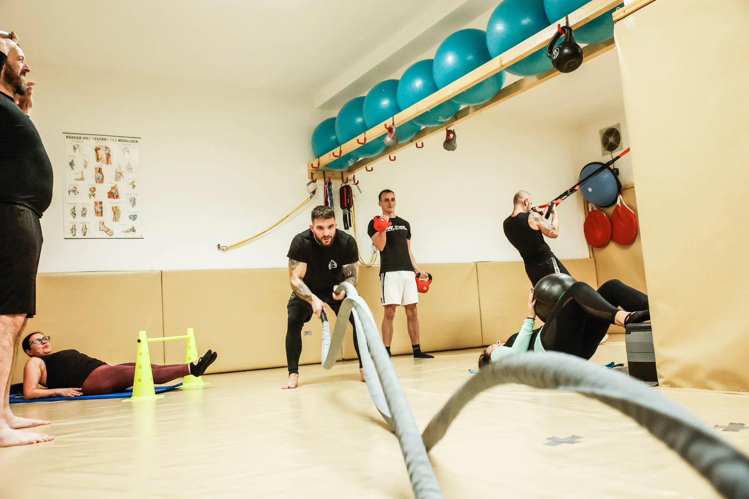 Skupina na kruhovém tréninku: muž v popředí pracuje se zátěžovými lany, ostatní cvičí na svých stanovištích u stěny.