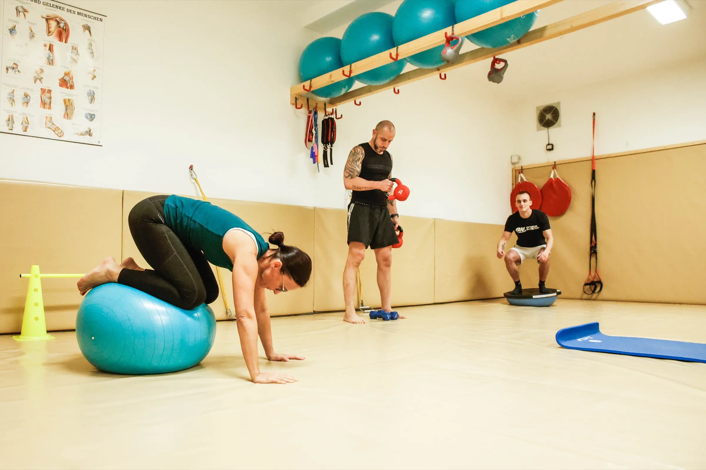 Kruhový trénink v tělocvičně: žena cvičí na gymnastickém míči, další dva účastníci pracují s jednoručkami a na balanční podložce.